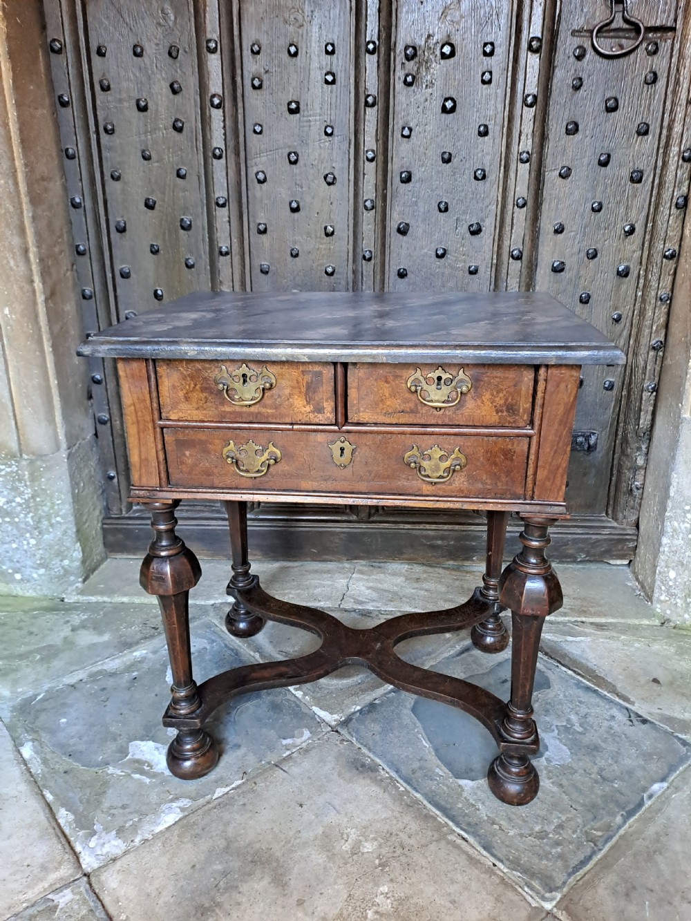A walnut side table featuring three drawers with brass handles, inverted cup turned legs, and a curved X-stretcher. View VI