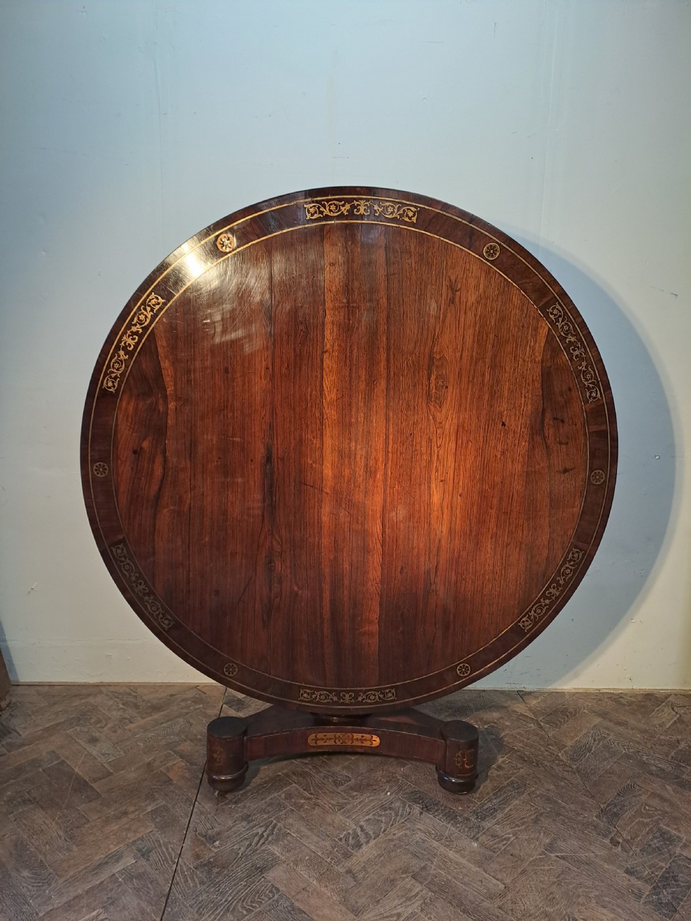 A Regency circular rosewood centre table shown tilted vertically, featuring floral brass marquetry inlay along the perimeter and base.