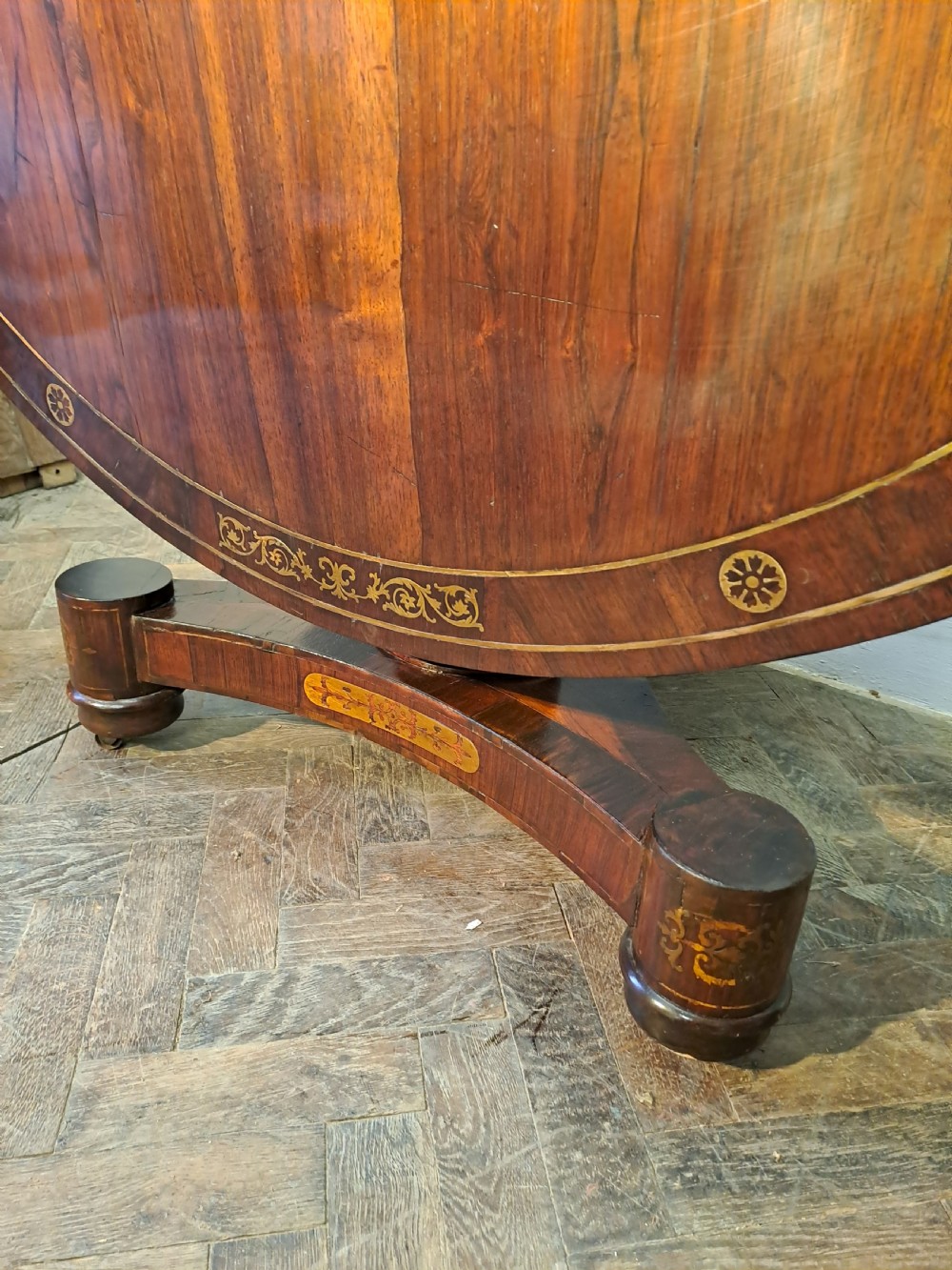 A Regency circular rosewood centre table shown tilted vertically, featuring floral brass marquetry inlay along the perimeter and base. View VI