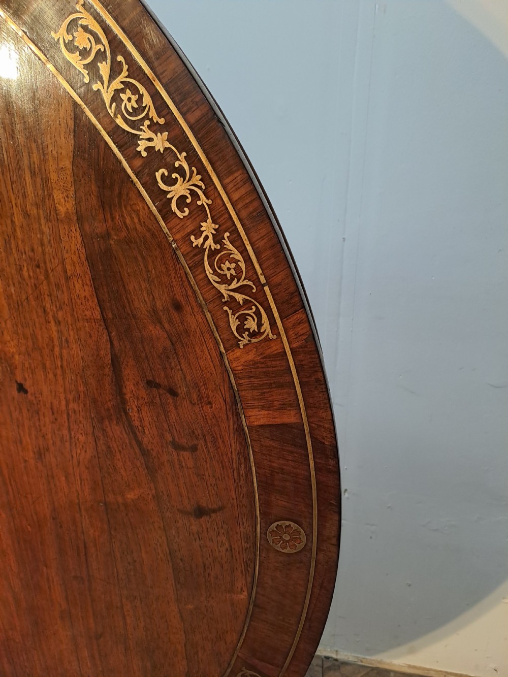 A Regency circular rosewood centre table shown tilted vertically, featuring floral brass marquetry inlay along the perimeter and base. View VIII