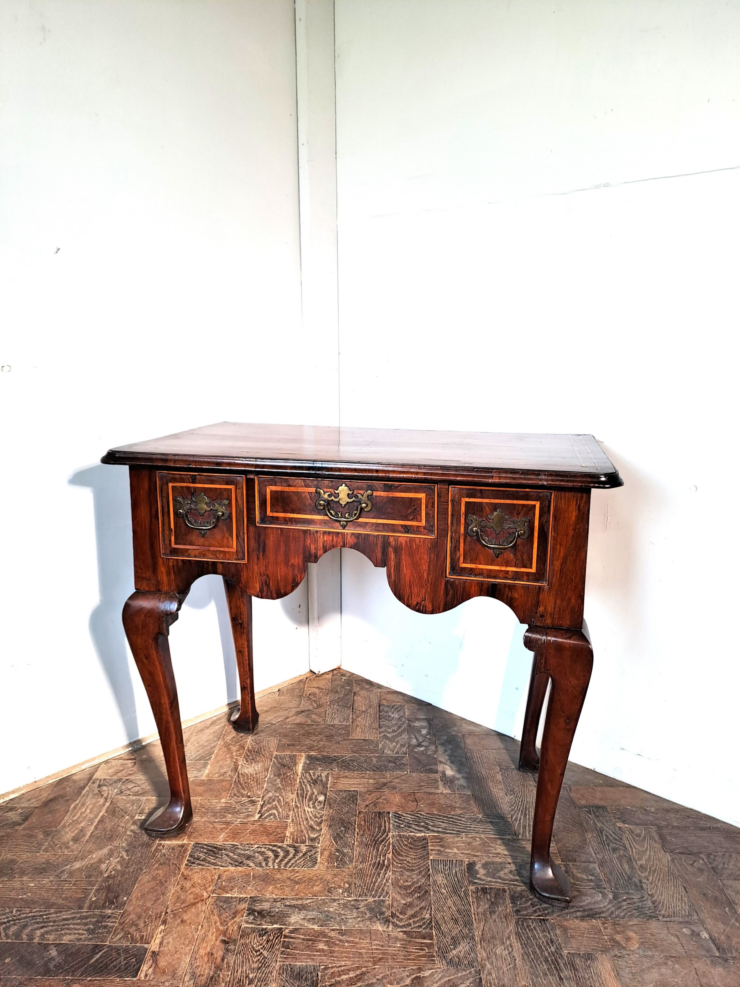 An early 18th-century walnut lowboy featuring three crossbanded drawers, brass handles, a shaped apron, and four cabriole legs.