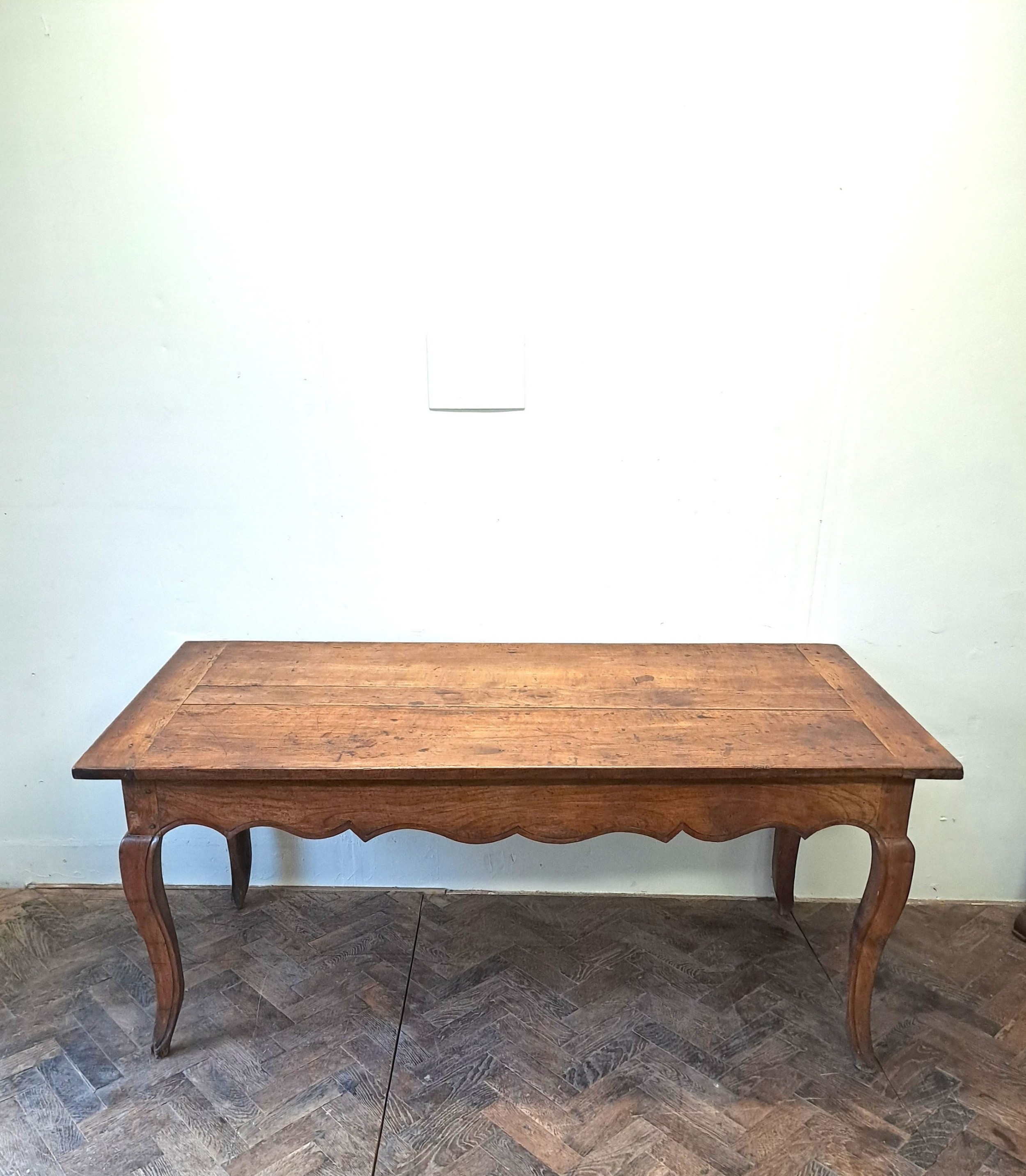 An early 19th-century French farmhouse kitchen table made of warm brown wood with a plank top, scalloped apron, and curved cabriole legs.