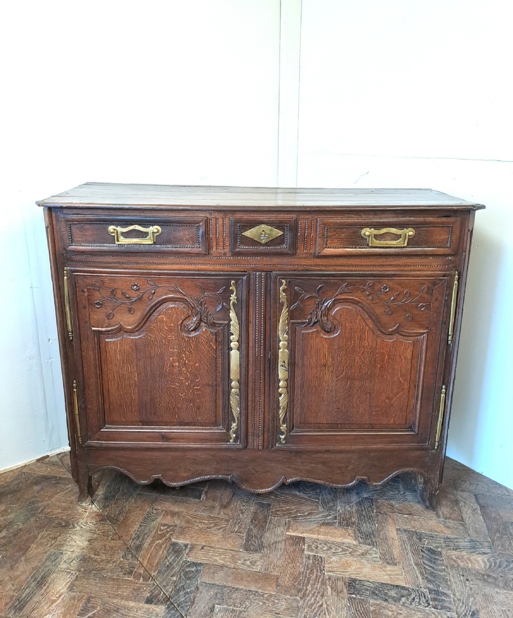 An antique French oak dresser base featuring carved foliate door panels, two top drawers, and decorative brass hardware.