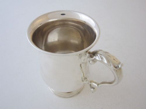fig 1. A plain sterling silver christening mug featuring a flared rim, baluster body, pedestal base, and a leaf-capped scroll handle.