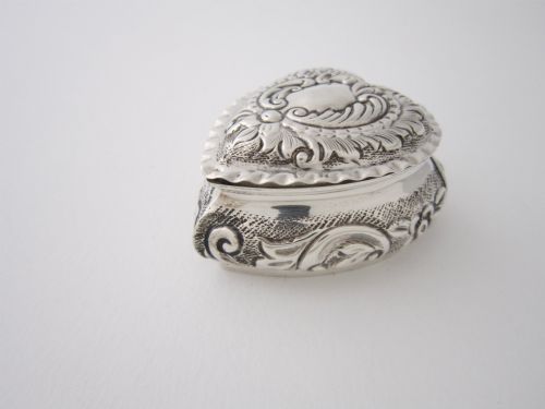 fig 1. A small heart-shaped sterling silver trinket box featuring intricate repousse scrollwork, floral motifs, a plain central cartouche, and a crimped rim.