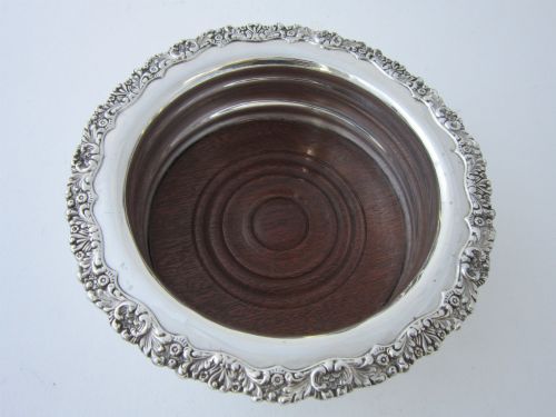 fig 1. A circular George V sterling silver wine coaster featuring an ornate floral scroll rim and a polished stepped body with a wood insert base.