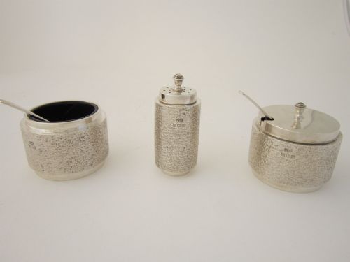 fig 2. A three-piece Elizabeth II sterling silver condiment set with textured bark-effect bodies, including a mustard pot, salt pot, pepper pot, and two small spoons, housed in a blue presentation case.