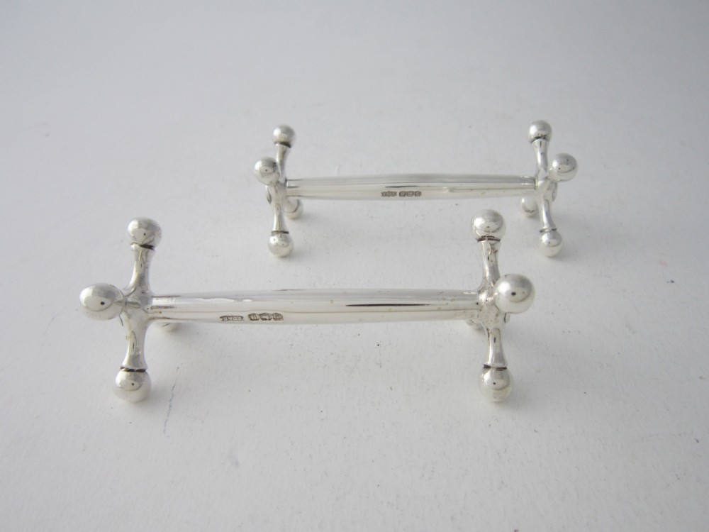 A pair of solid sterling silver knife rests featuring crossed X-shaped end supports with spherical terminals joined by a horizontal central bar. View III