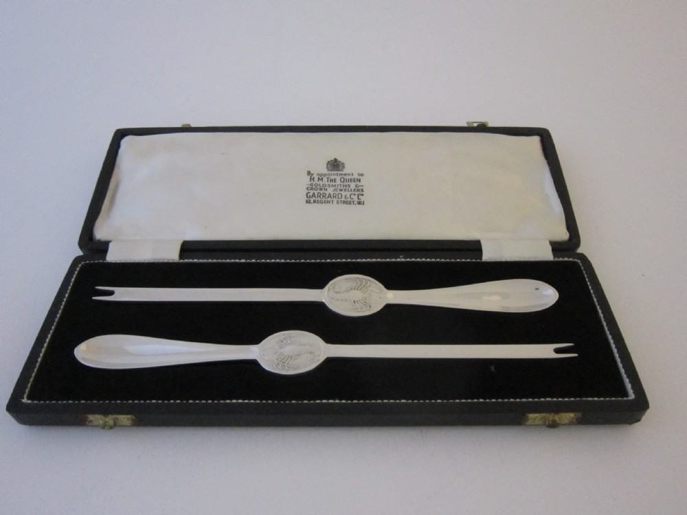 A pair of sterling silver lobster picks with embossed central medallions displayed inside an open black velvet-lined box.