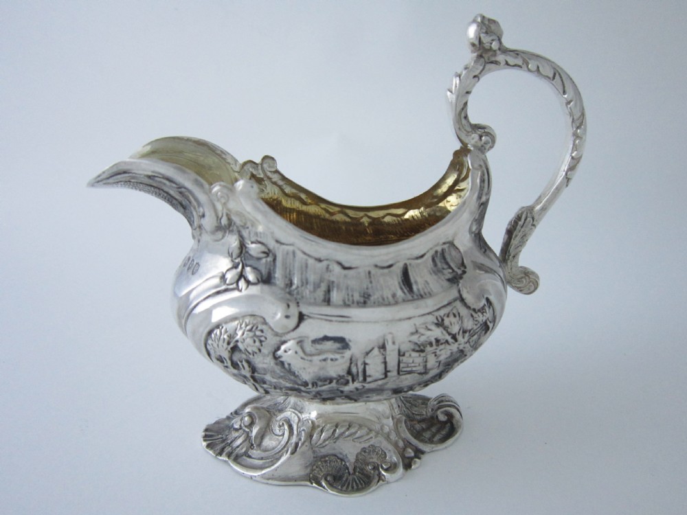 Victorian sterling silver cream jug with detailed repousse pastoral scenes, a high scroll handle, a gilt interior, and a cast foot.