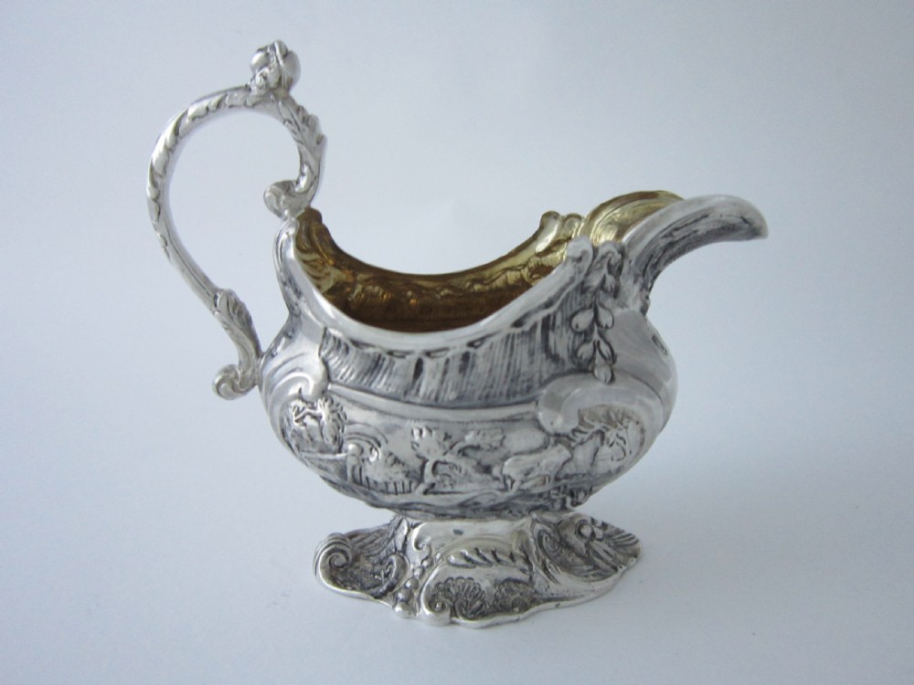 Victorian sterling silver cream jug with detailed repousse pastoral scenes, a high scroll handle, a gilt interior, and a cast foot. View III