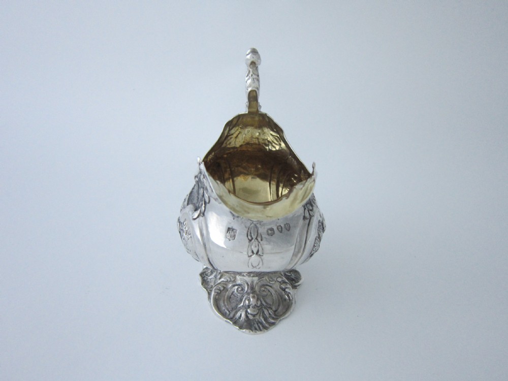 Victorian sterling silver cream jug with detailed repousse pastoral scenes, a high scroll handle, a gilt interior, and a cast foot. View V