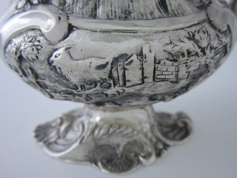 Victorian sterling silver cream jug with detailed repousse pastoral scenes, a high scroll handle, a gilt interior, and a cast foot. View VI