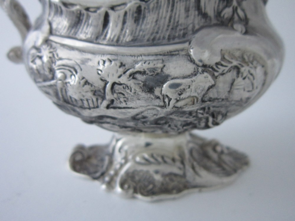Victorian sterling silver cream jug with detailed repousse pastoral scenes, a high scroll handle, a gilt interior, and a cast foot. View VII
