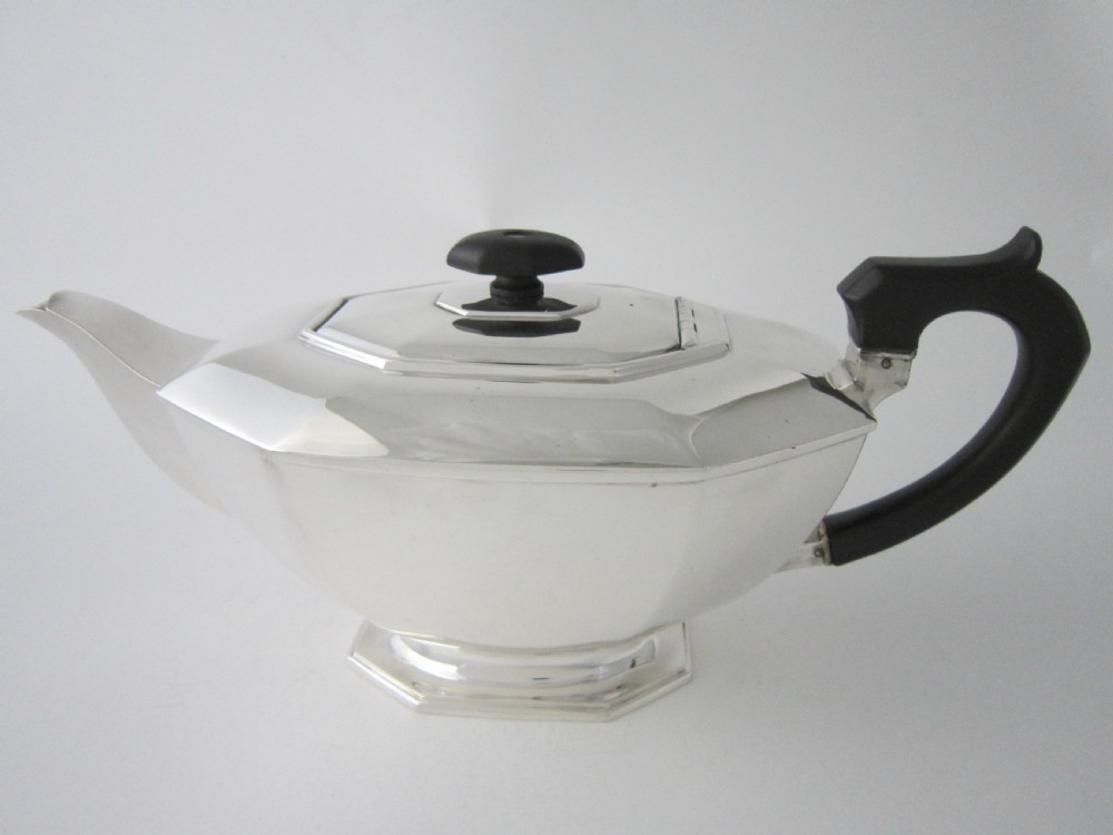 An octagonal sterling silver teapot featuring a carved black wooden handle and matching black lid finial, resting on a faceted pedestal base.