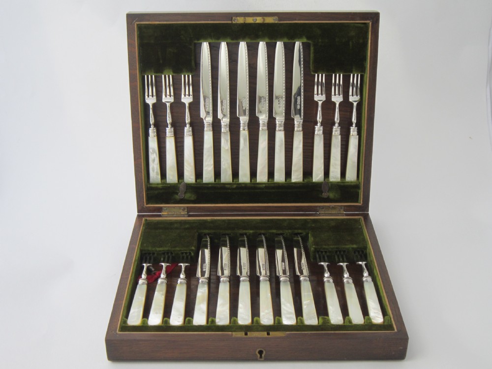 An open fitted wooden canteen lined with green velvet, containing a set of sterling silver dessert knives and forks with carved mother-of-pearl handles.