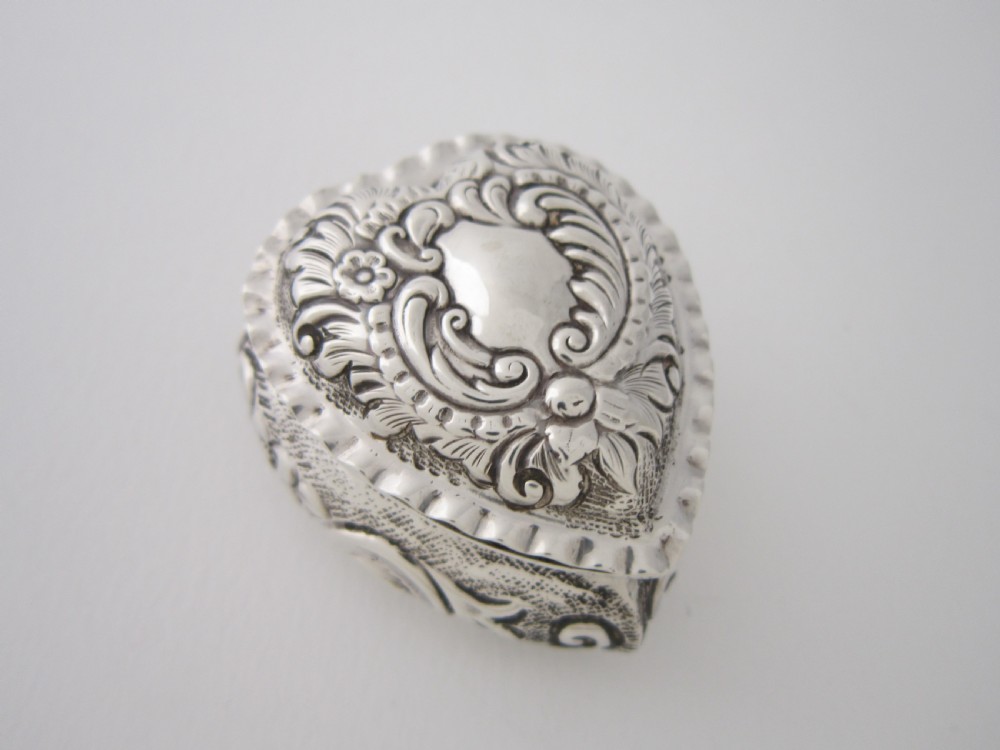 A heart-shaped sterling silver trinket box with detailed embossed foliate scrollwork and a scalloped rim.