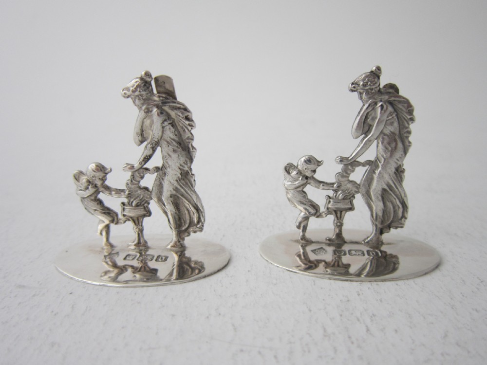 A pair of Edwardian sterling silver place card holders depicting classical figures and cherubs on circular hallmarked bases.