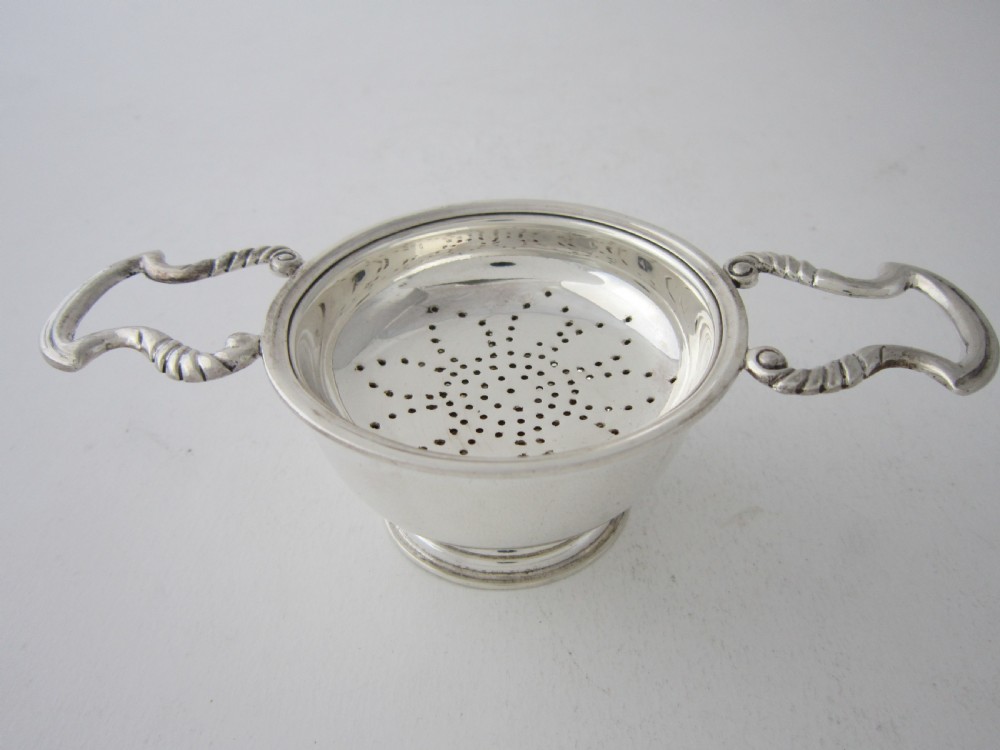 A solid sterling silver double-handled tea strainer with a pierced star bowl pattern and scrolled side handles.