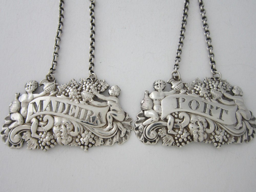 A pair of Scottish George III sterling silver decanter labels engraved with Madeira and Port, featuring relief motifs of cherubs, grapes, and scrollwork on hanging chains.