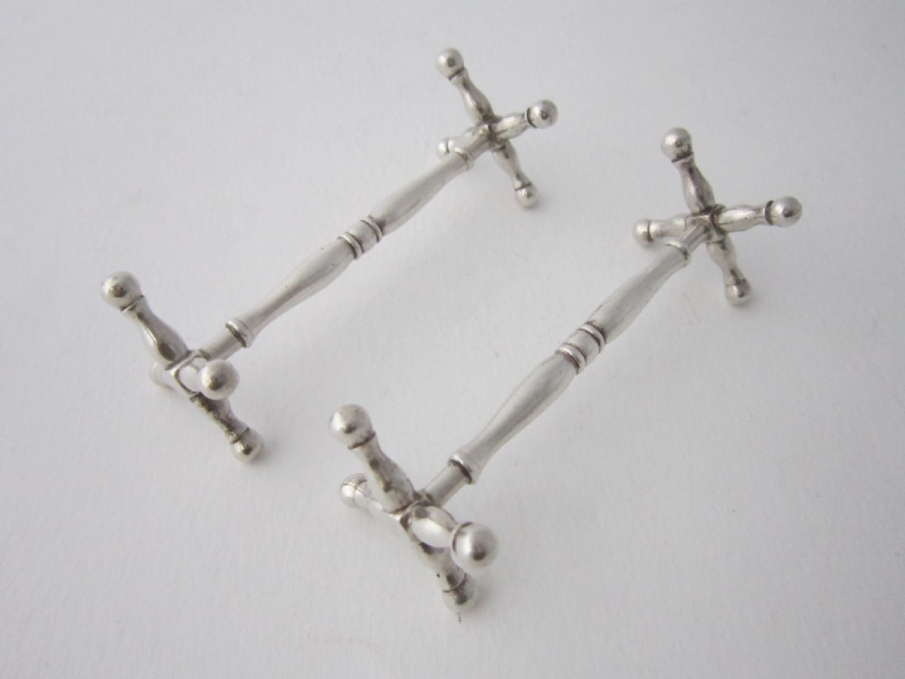 A pair of antique sterling silver knife rests featuring turned central bars and cross-shaped end supports with ball feet.