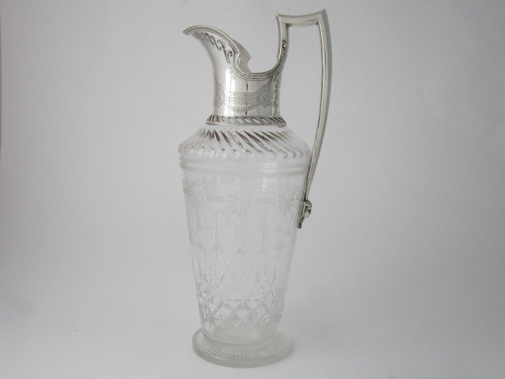 An antique Victorian claret jug featuring a clear etched glass body with cut geometric patterns and an engraved sterling silver collar and handle.