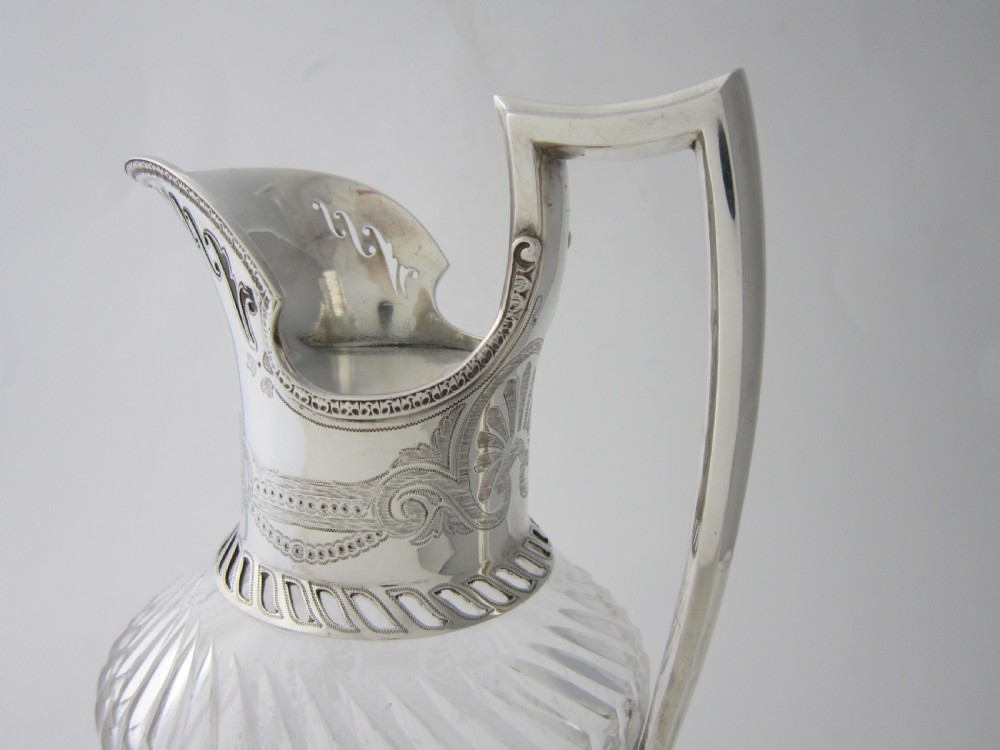 An antique Victorian claret jug featuring a clear etched glass body with cut geometric patterns and an engraved sterling silver collar and handle. View X