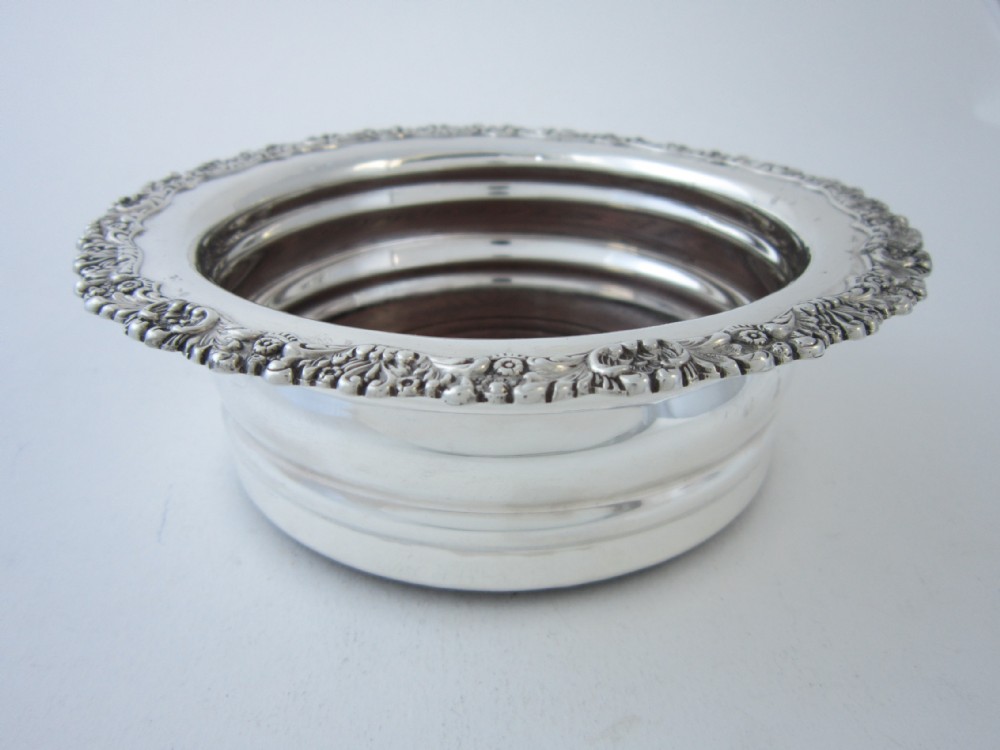 A circular George V sterling silver wine coaster featuring an ornate floral scroll rim and a deep turned wood interior base.