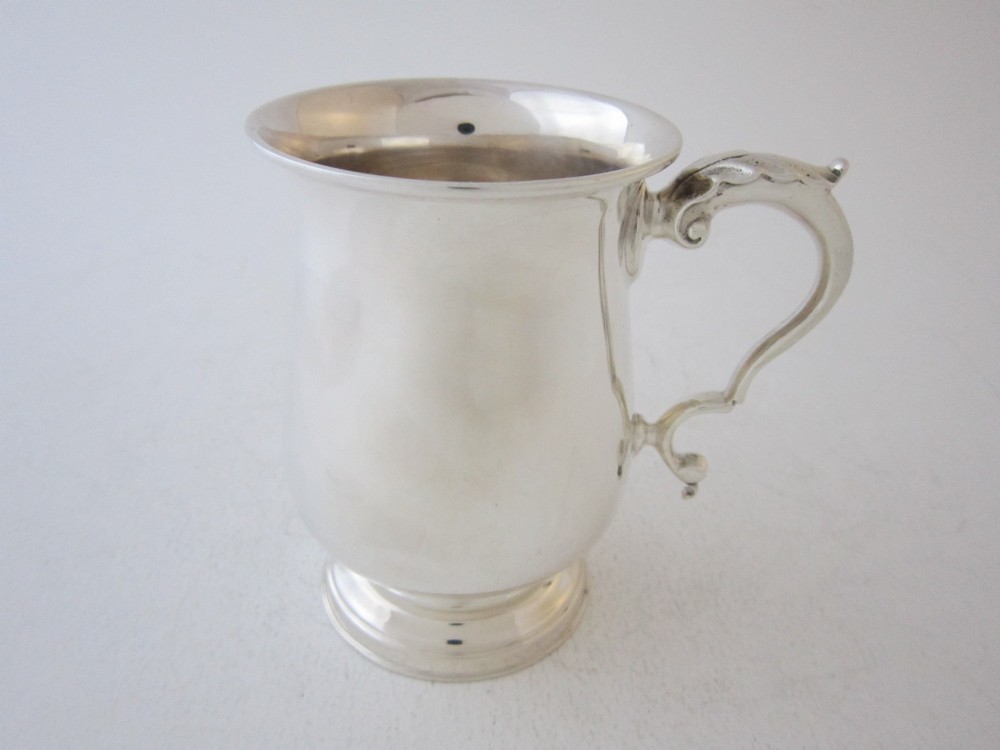 A plain sterling silver christening mug featuring a flared rim, baluster body, pedestal base, and a leaf-capped scroll handle.