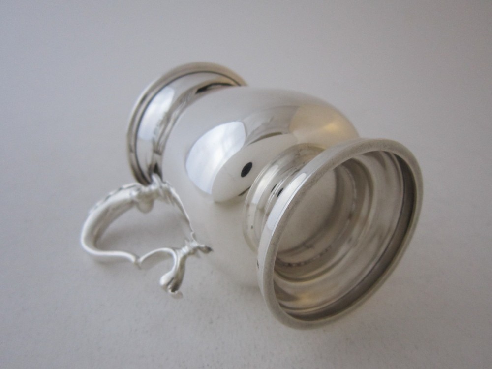 A plain sterling silver christening mug featuring a flared rim, baluster body, pedestal base, and a leaf-capped scroll handle. View IV