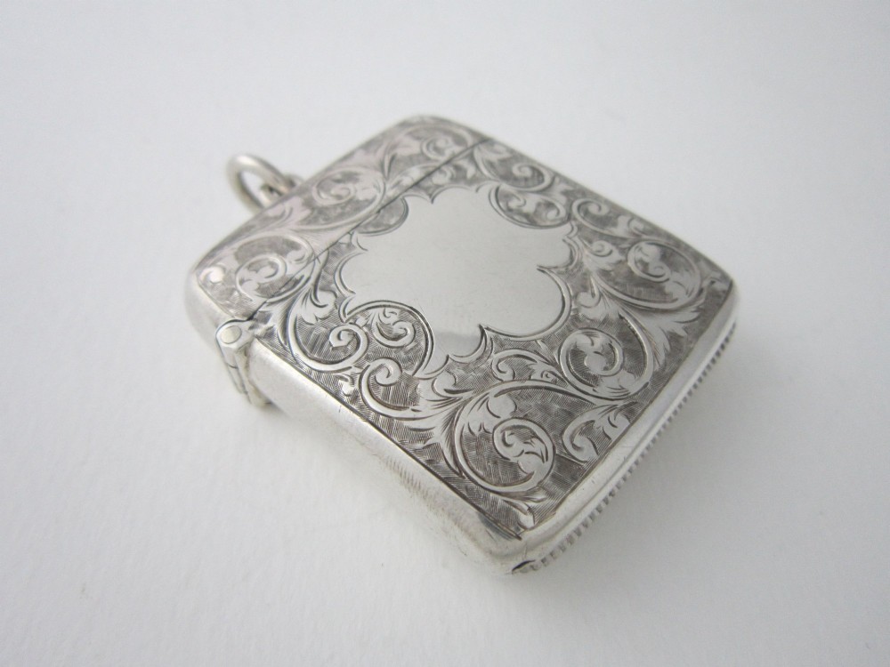 An antique sterling silver vesta case featuring hand-engraved foliate scrollwork, a vacant shield cartouche, and an attached suspension ring.