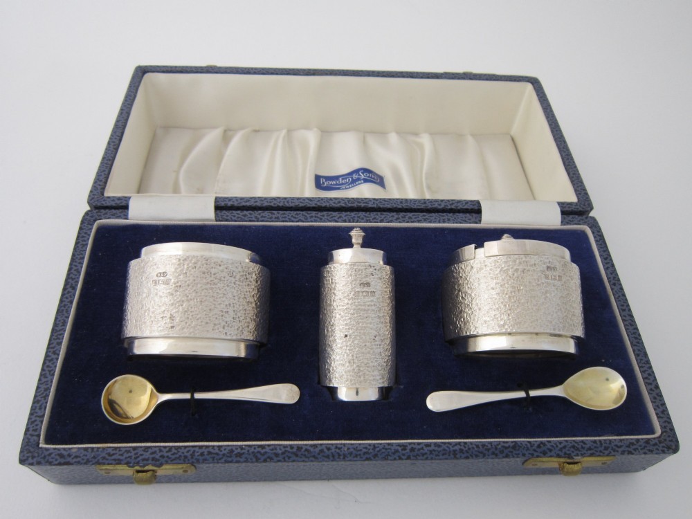 A three-piece sterling silver condiment set with textured bark-effect surfaces including a salt cellar, mustard pot, pepper shaker, and two spoons in a blue fitted case.