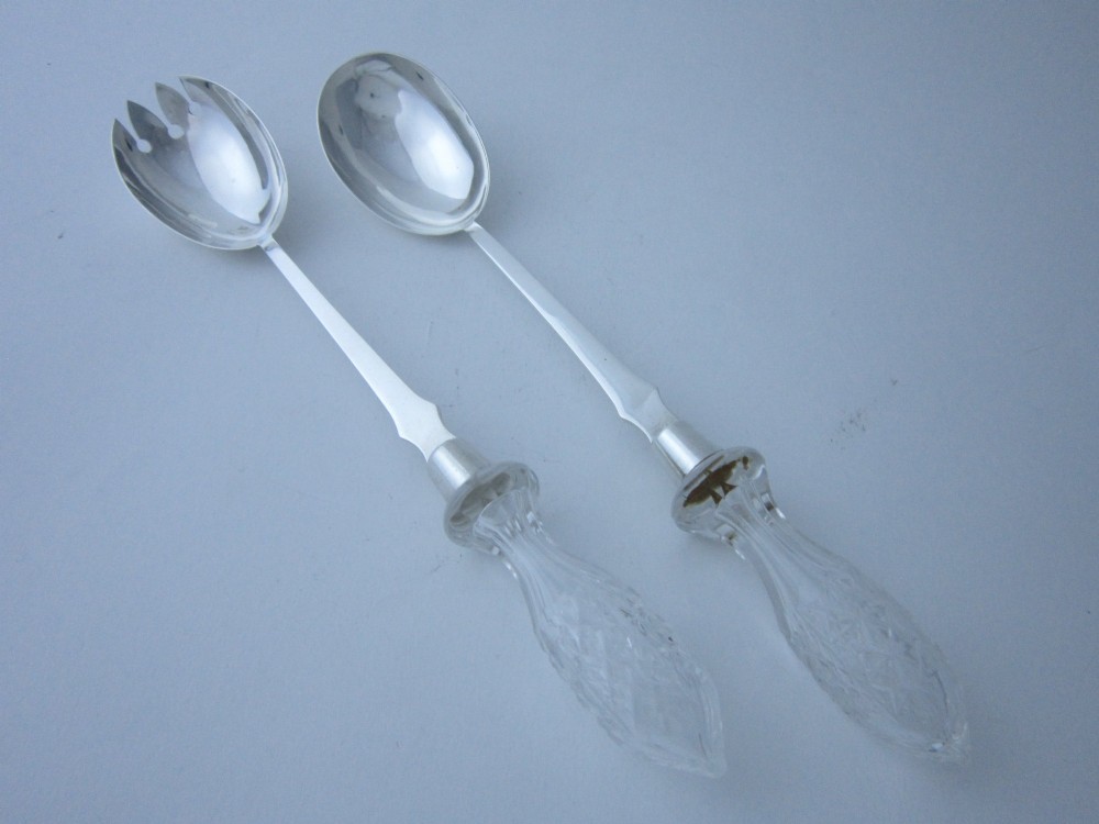 A pair of Edwardian sterling silver salad servers featuring faceted cut glass handles and rat tail detailing on the bowls.