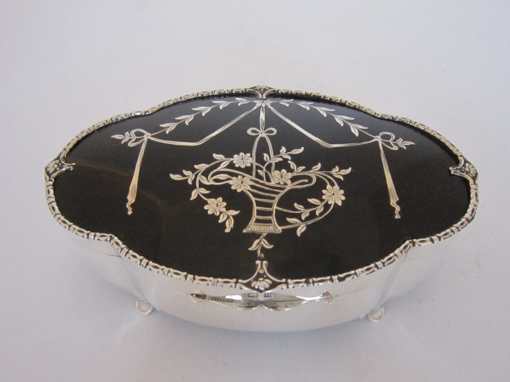 A shaped sterling silver trinket box featuring a dark tortoiseshell lid inlaid with a silver flower basket and ribbon swags, resting on four small feet.