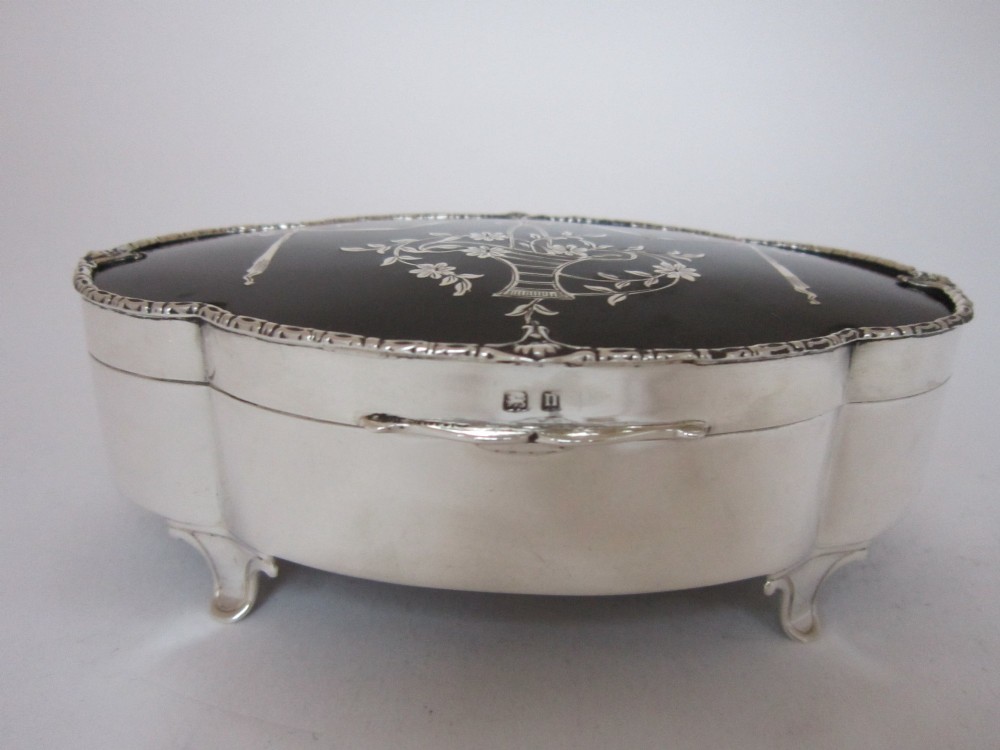 A shaped sterling silver trinket box featuring a dark tortoiseshell lid inlaid with a silver flower basket and ribbon swags, resting on four small feet. View II
