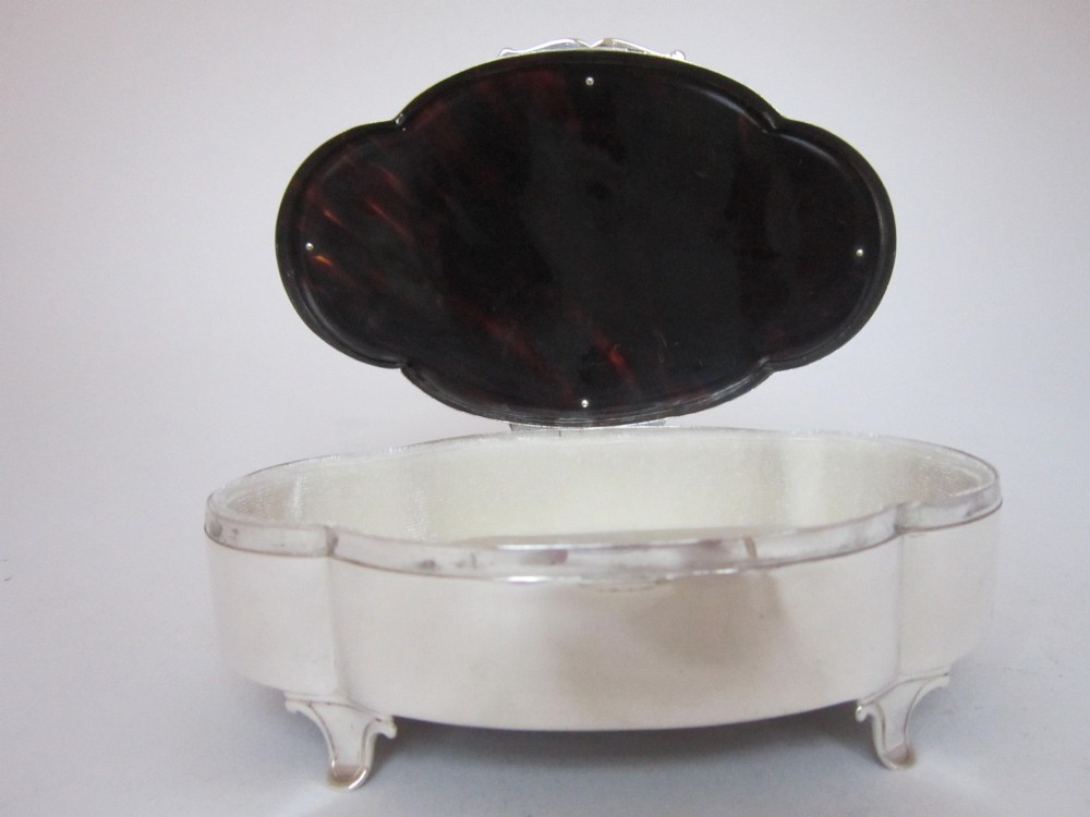A shaped sterling silver trinket box featuring a dark tortoiseshell lid inlaid with a silver flower basket and ribbon swags, resting on four small feet. View III