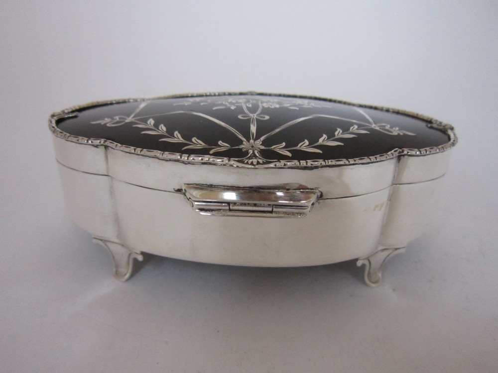 A shaped sterling silver trinket box featuring a dark tortoiseshell lid inlaid with a silver flower basket and ribbon swags, resting on four small feet. View V