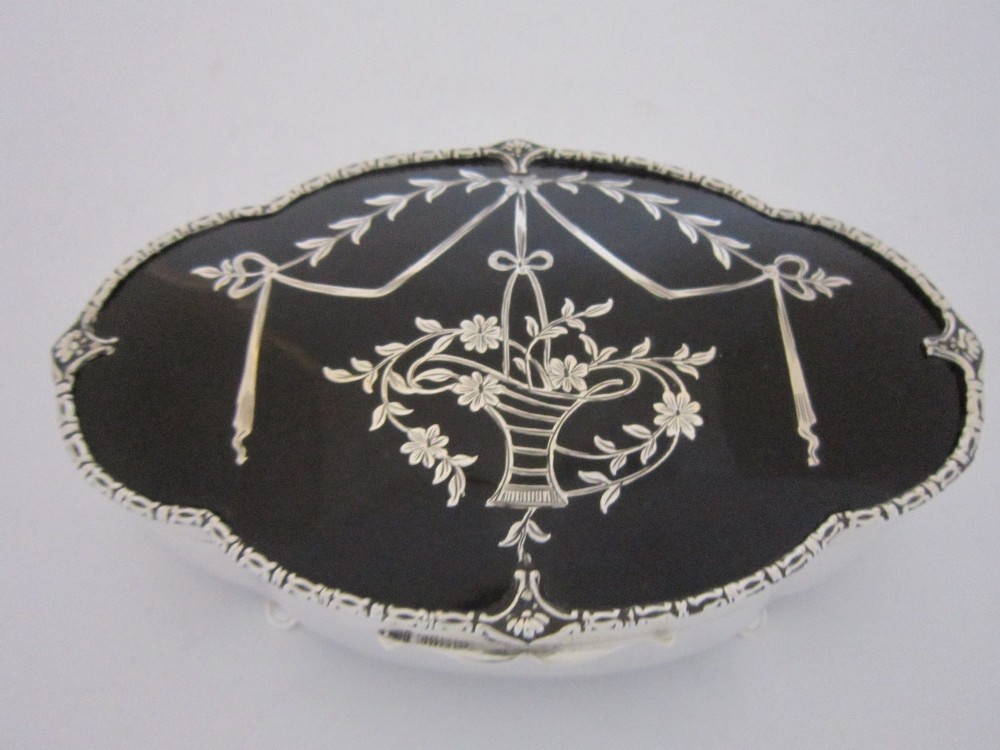 A shaped sterling silver trinket box featuring a dark tortoiseshell lid inlaid with a silver flower basket and ribbon swags, resting on four small feet. View VI