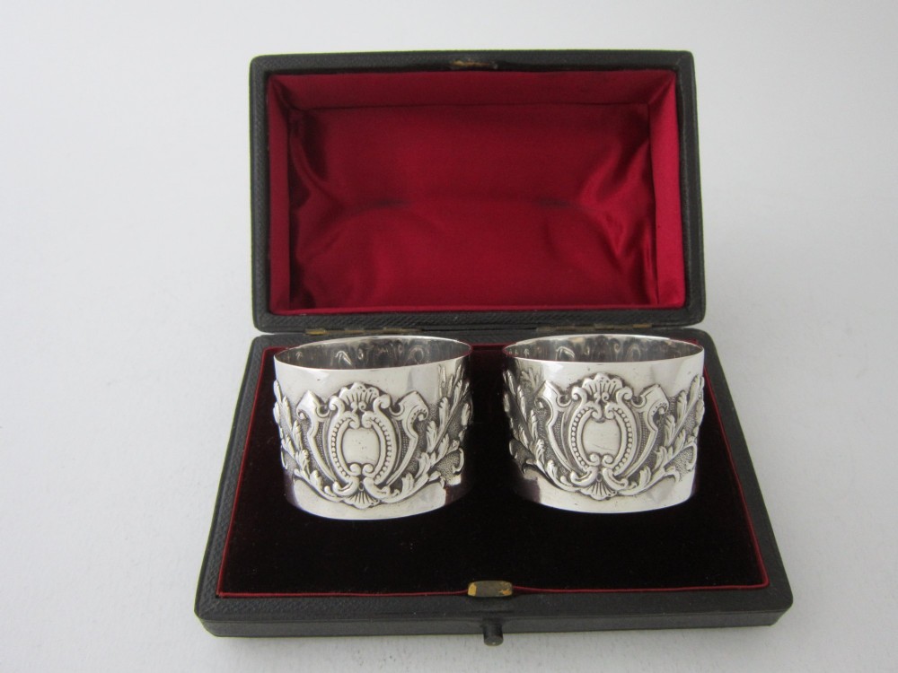 A pair of ornate Victorian sterling silver napkin rings featuring embossed acanthus leaves and cartouches inside a velvet-lined black presentation box.