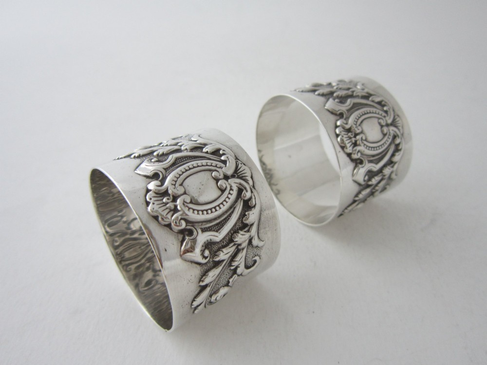 A pair of ornate Victorian sterling silver napkin rings featuring embossed acanthus leaves and cartouches inside a velvet-lined black presentation box. View V
