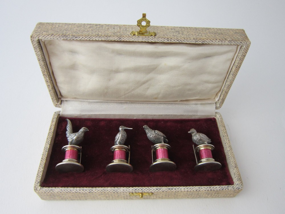 A set of four Edwardian sterling silver place card holders featuring pink guilloche enamel columns topped with sculpted game birds inside a fitted presentation box.