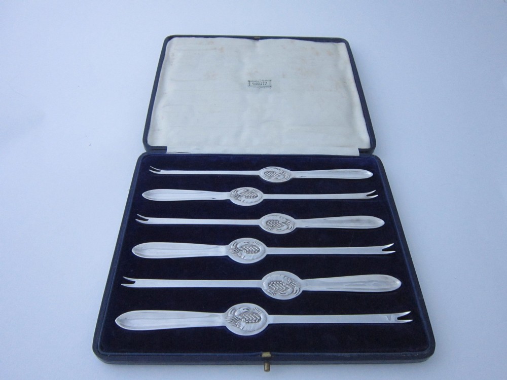 A set of six George V sterling silver lobster picks featuring central embossed lobster medallions, presented inside an open navy blue fitted velvet display case.