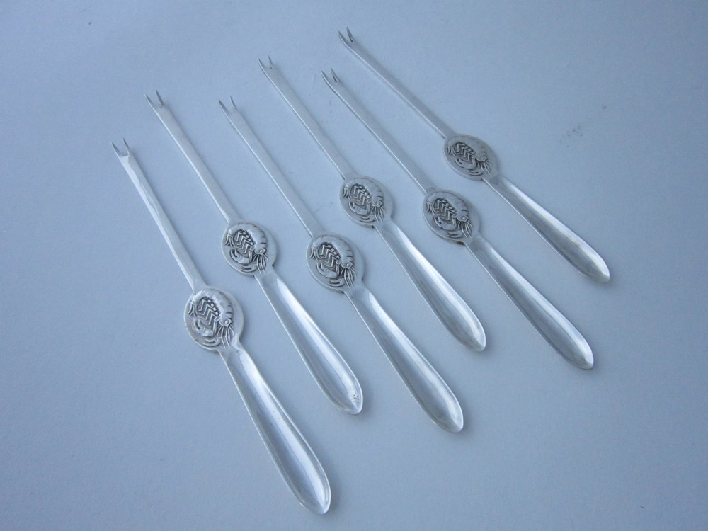 A set of six George V sterling silver lobster picks featuring central embossed lobster medallions, presented inside an open navy blue fitted velvet display case. View III