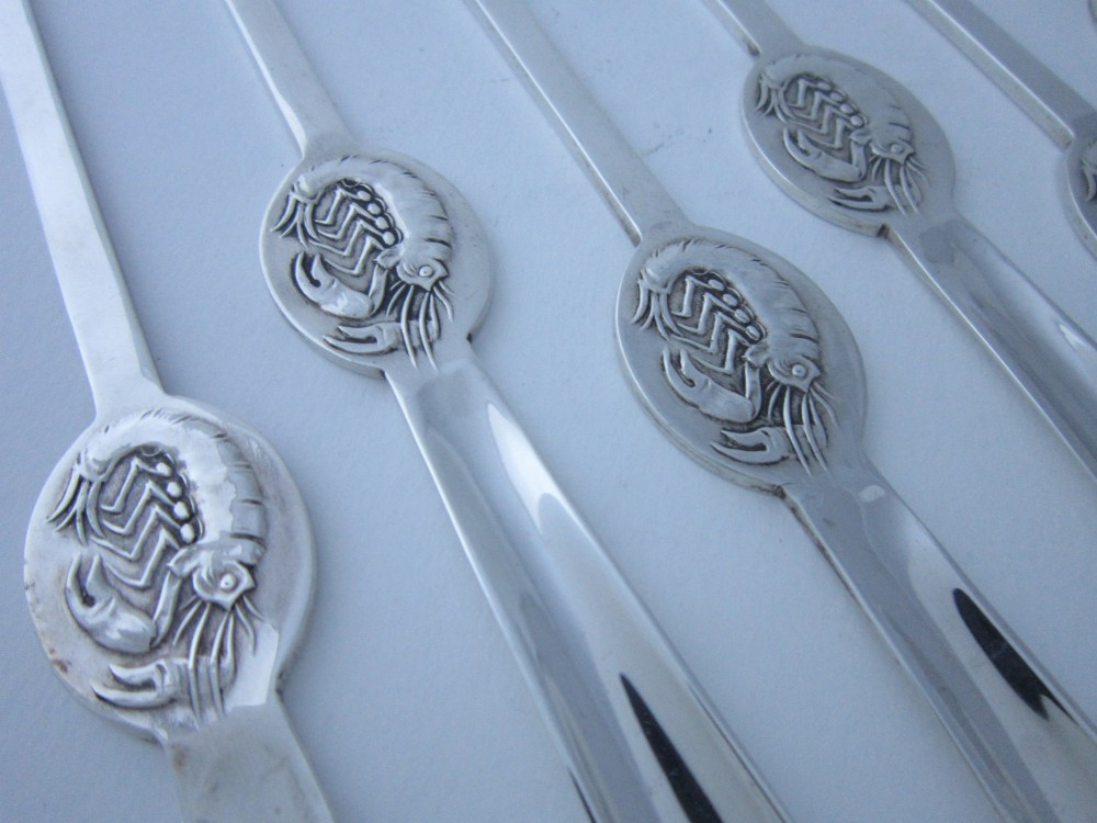 A set of six George V sterling silver lobster picks featuring central embossed lobster medallions, presented inside an open navy blue fitted velvet display case. View V