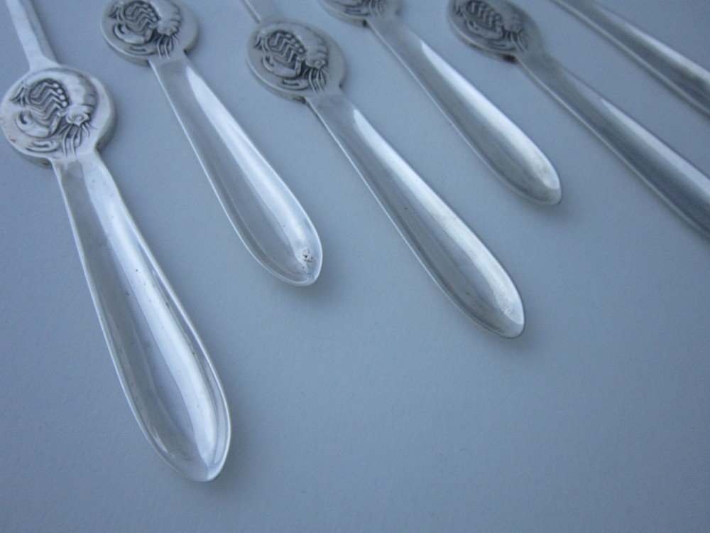 A set of six George V sterling silver lobster picks featuring central embossed lobster medallions, presented inside an open navy blue fitted velvet display case. View VI