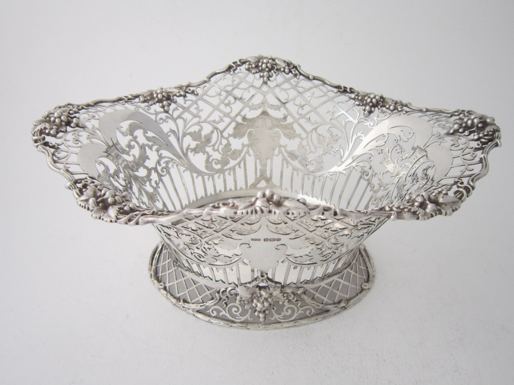An antique George V sterling silver basket featuring intricate pierced lattice metalwork, scrolled grapevine borders, and visible hallmarks. View II