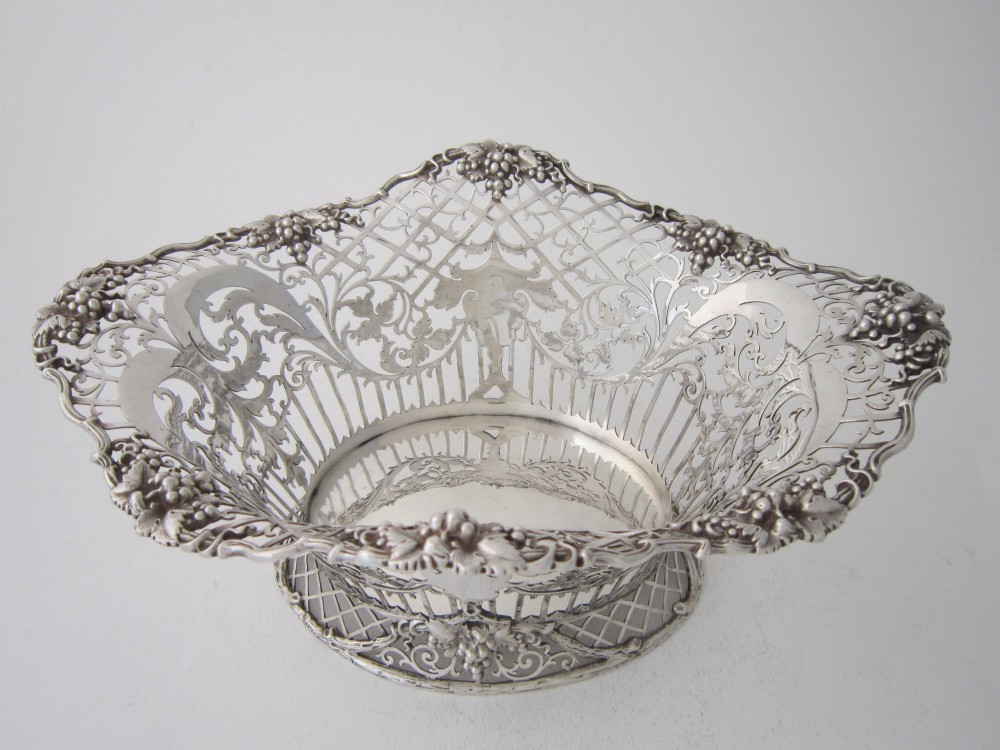An antique George V sterling silver basket featuring intricate pierced lattice metalwork, scrolled grapevine borders, and visible hallmarks. View III