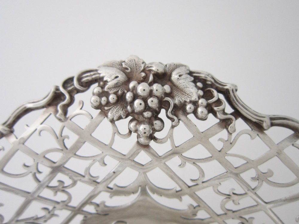An antique George V sterling silver basket featuring intricate pierced lattice metalwork, scrolled grapevine borders, and visible hallmarks. View IV