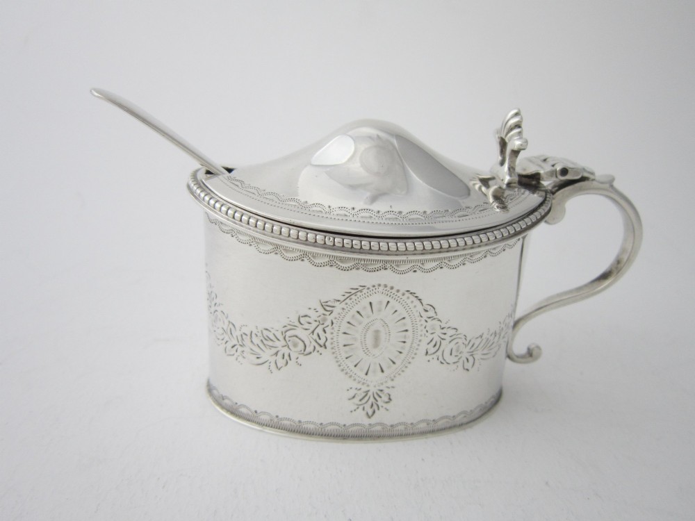 An oval sterling silver mustard pot featuring bright-cut engraved patterns, a beaded rim, a scroll handle, and a matching spoon.