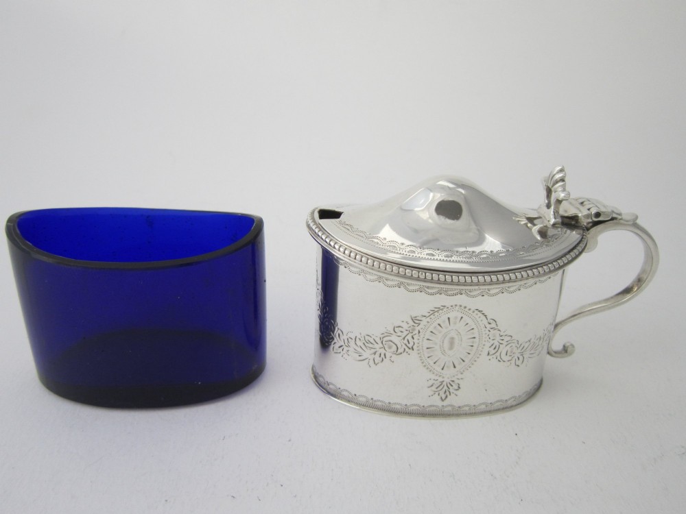 An oval sterling silver mustard pot featuring bright-cut engraved patterns, a beaded rim, a scroll handle, and a matching spoon. View X